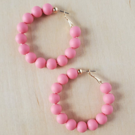 large bright pink rubber beads strung onto gold hoop earrings, resting on a natural wood table
