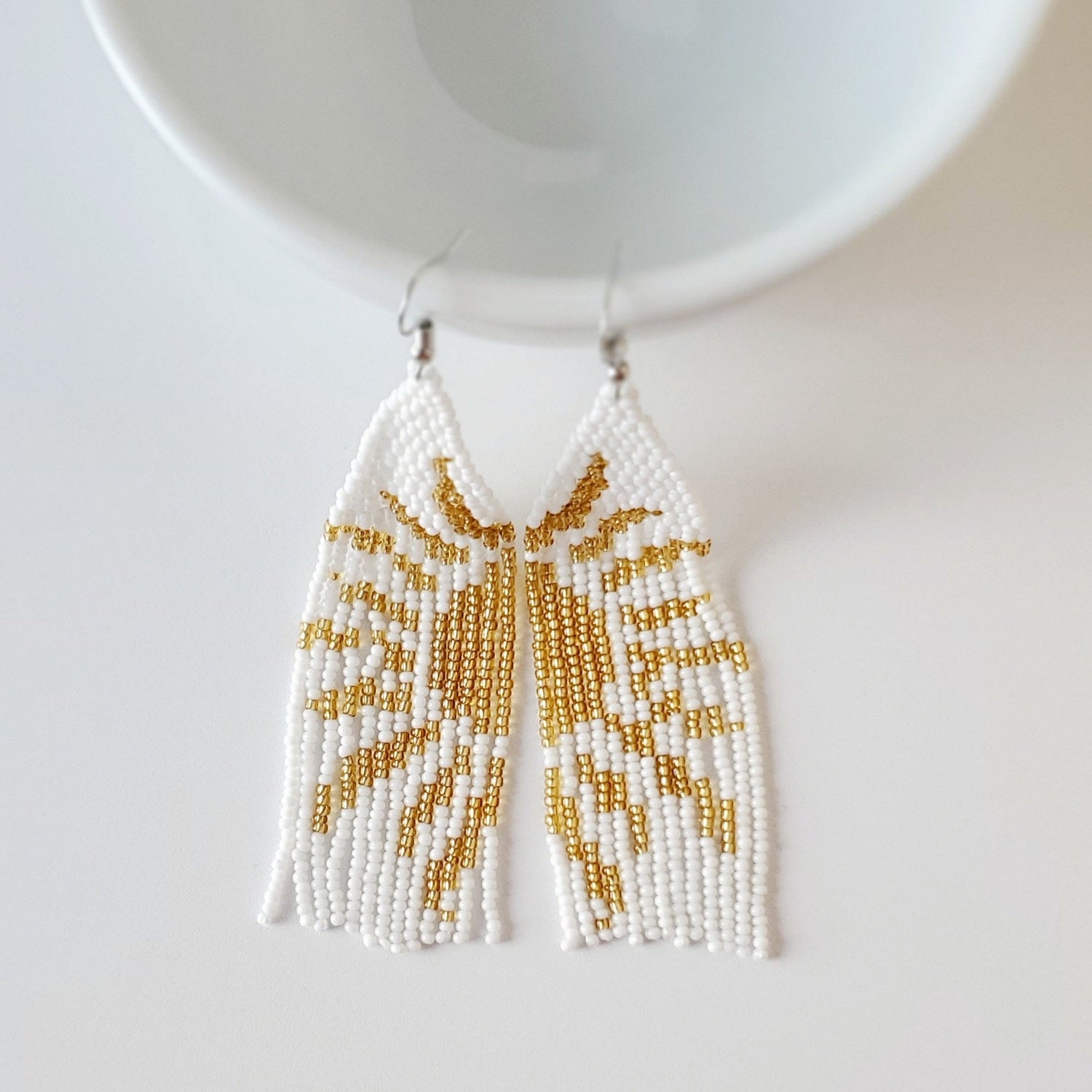 top middle is a shiny white ceramic bowl sitting on a white table. the bowl has 2 earrings with silver hooks hooked on to the edge of it. The earrings are 4.75" long with white seed beads and golden seed beads woven into a sun 