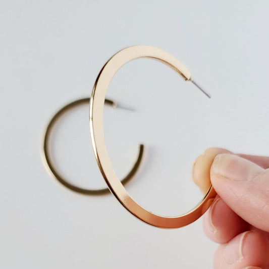 Medium sized, shiny gold flat hoop earring being held up. The other hoop sits in the background on a white table