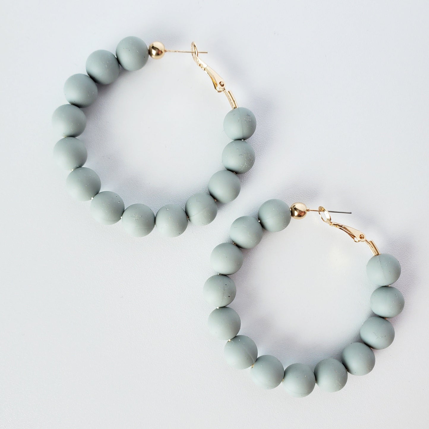 Sage colored rubber beads on a 2.25 inch gold hoop earring, sitting on a white table. the earrings are hinge back