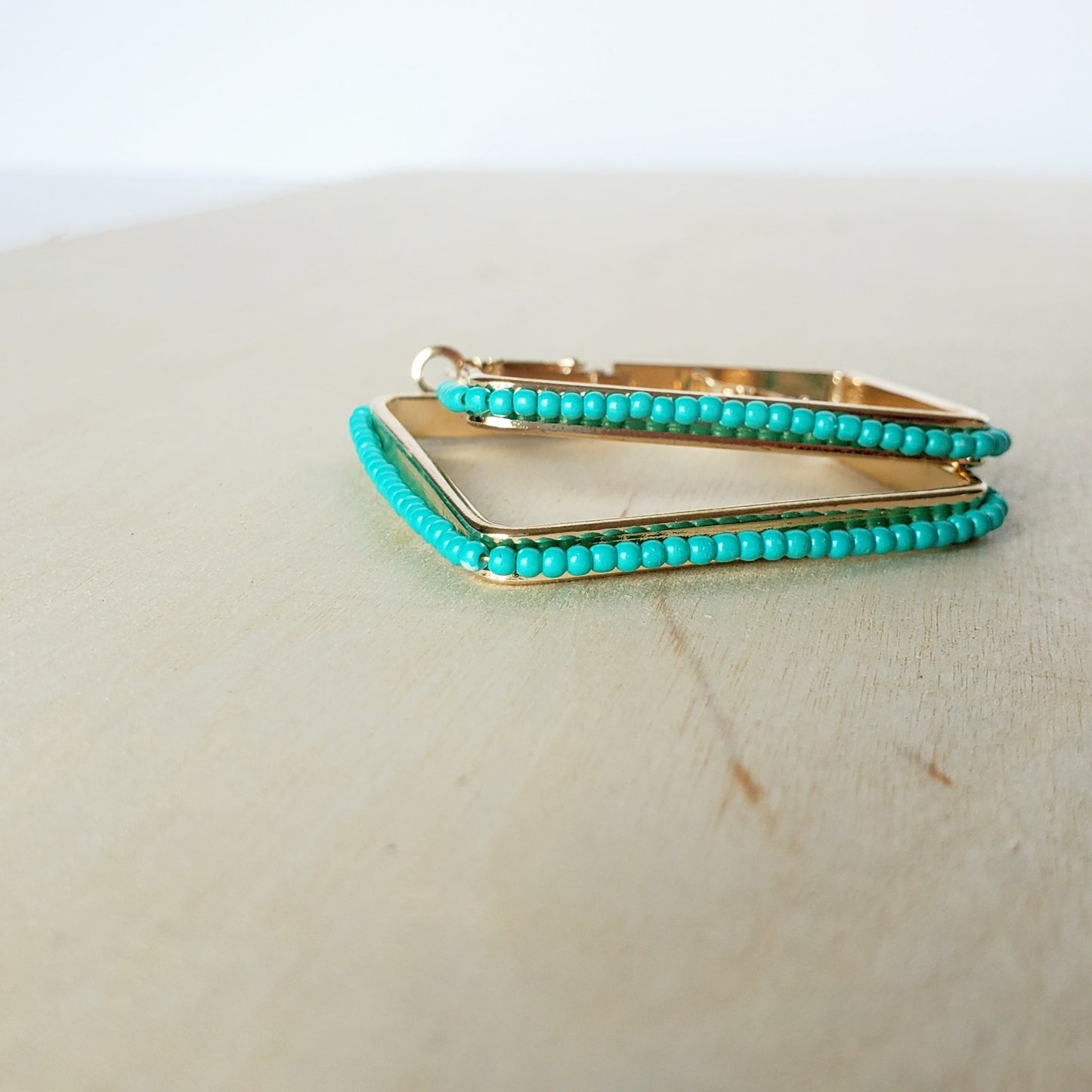 a pair of gold earrings with turquoise seed beads wrapped around the 2 inch square earrings, sit stacked on a light wood surface against a white wall