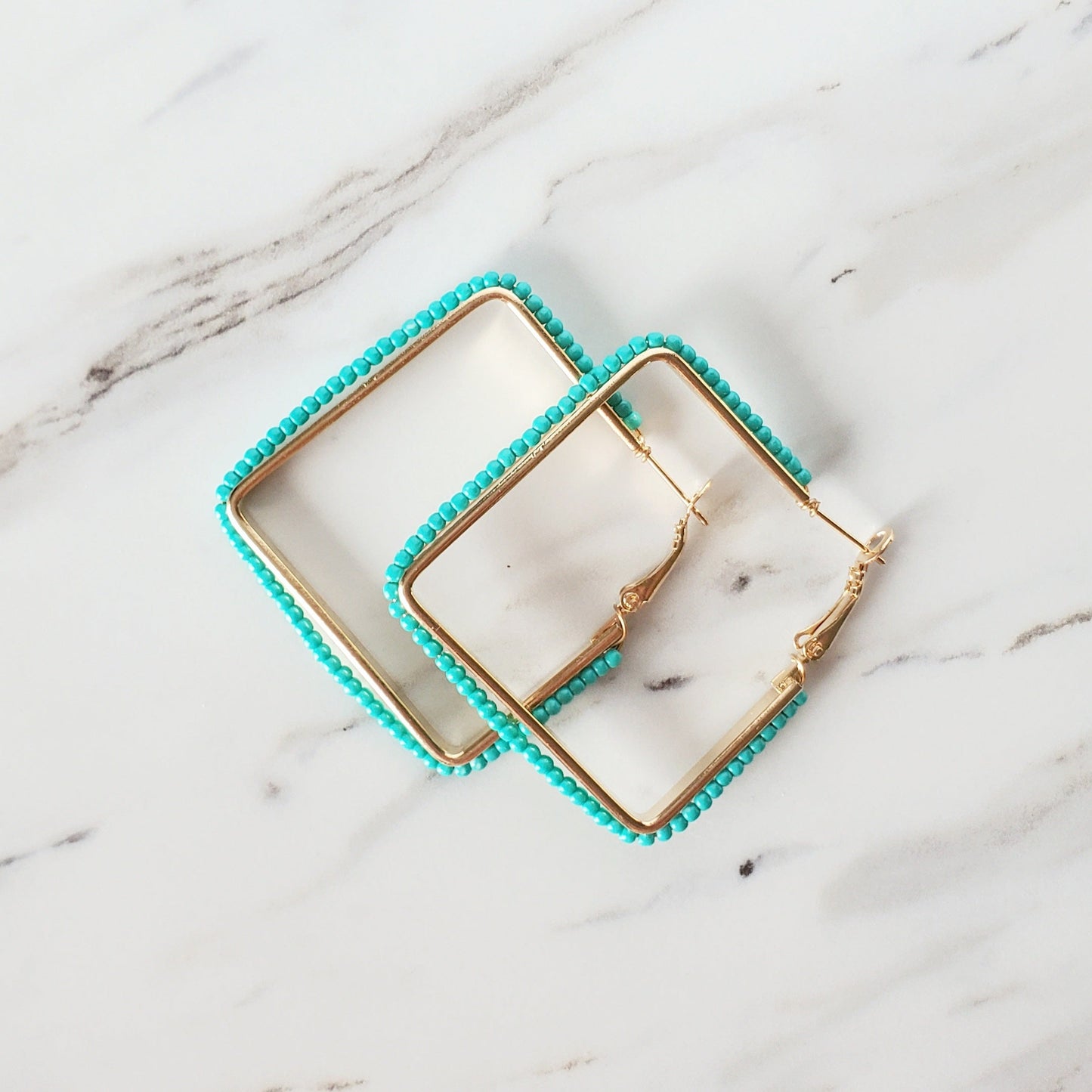 white and gray marbled table with a pair of 2 inch square, hinge back, gold metal earrings with turquoise seed beads wrapped around the metal in a straight line. the earrings are overlapping