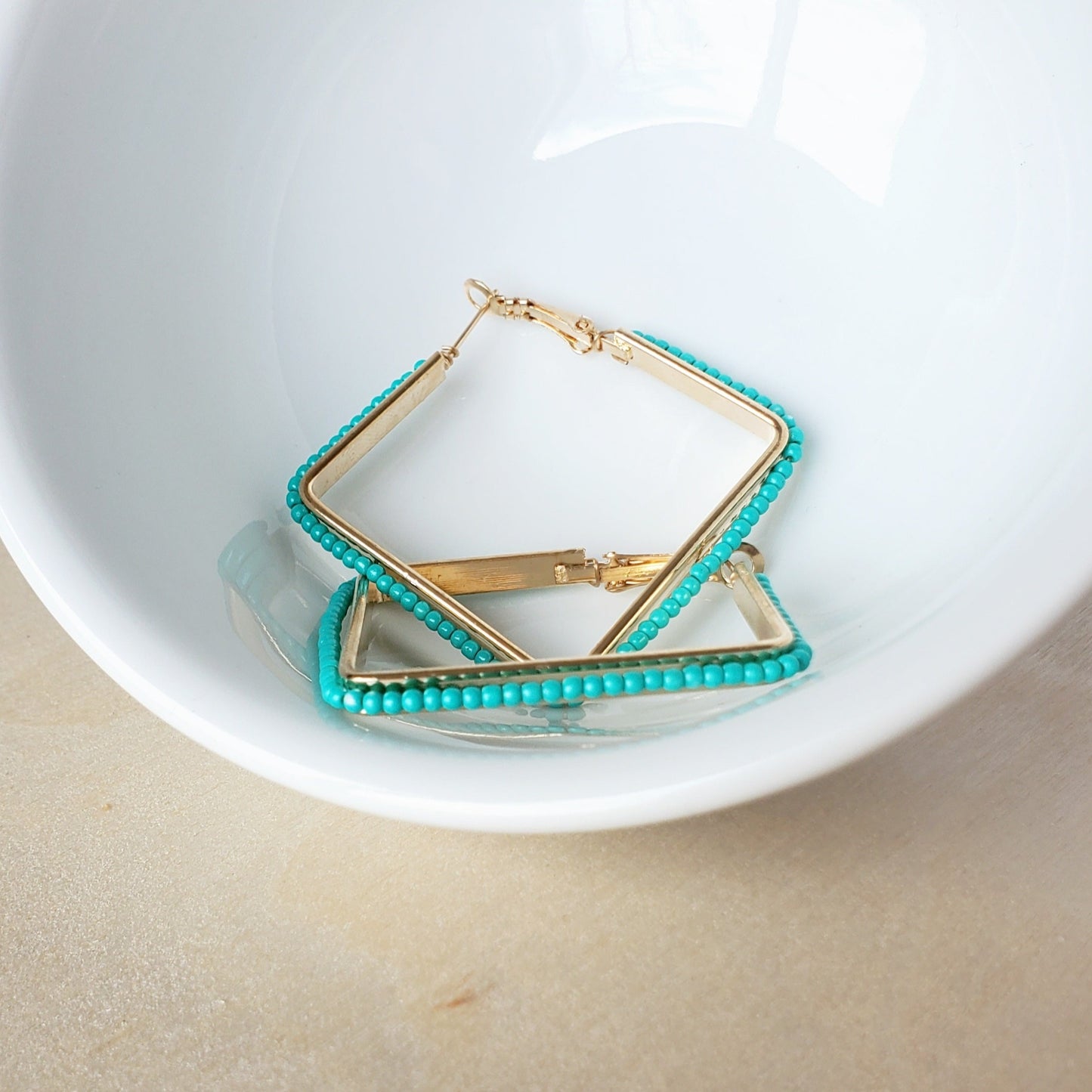 A shiny white ceramic bowl sits on a light wooden surface. in the bowl lays a pair of 2 inch square hinge back earrings. the earrings are made of gold metal wrapped with turquoise rice beads